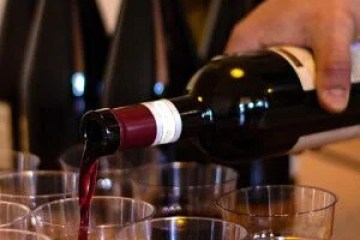 Red wine being poured into clear plastic cups, with wine bottles in the background.