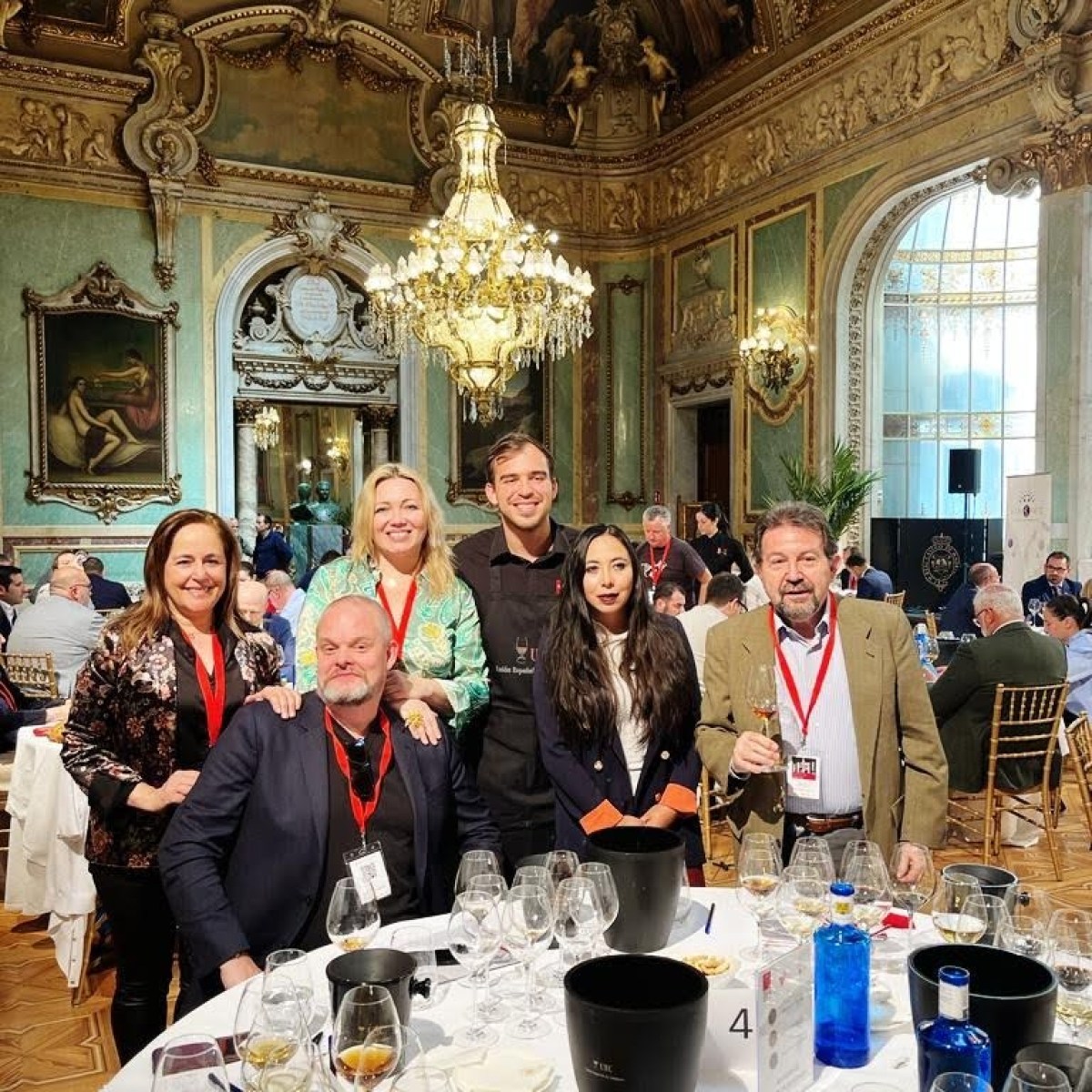 Group of people smiling around a table in an ornate room with chandeliers.